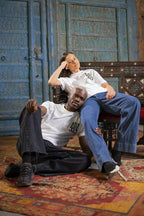 Two individuals sitting on a floor against a wooden backdrop, wearing 555 opulence white t-shirts with red text and designs.